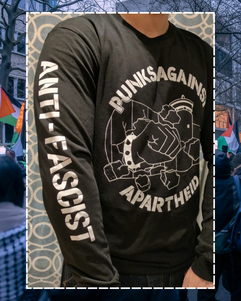 A photo of an individual facing forwards from the neck downards, showing the PAA logo (a fist with a spikey bracelet punching through the apartheid wall) & one arm which says ANTI-FASCIST.