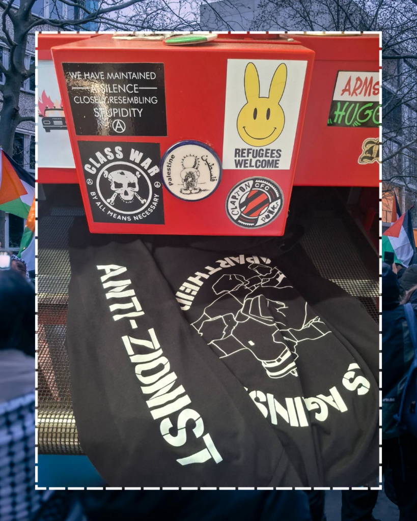 A machine which is printing designs on T-shirts, showing the PAA logo (a fist with a spikey bracelet punching through the apartheid wall) & one arm which says ANTI-ZIONIST. The machine has stickers: 'CLASS WAR BY ALL MEANS NECESSARY' with a skull in the centre, Clapton Punks, a smiling rabbit 'Refugees Welcome', a Handhala that says Palestine/فلسطين, and a sticker which says WE HAVE MAINTAINED A SILENCE CLOSELY RESEMBLING STUPIDITY.